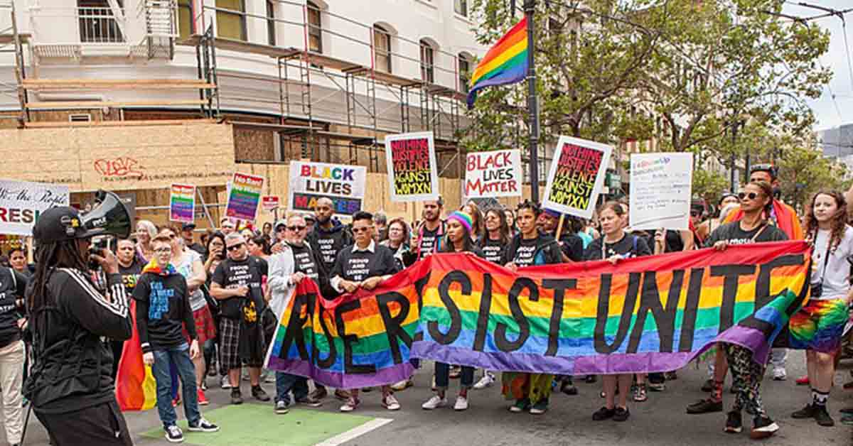 LGBTQ rally with people holding up "Rise Resist Unite" banner, a rainbow flag waving in the background