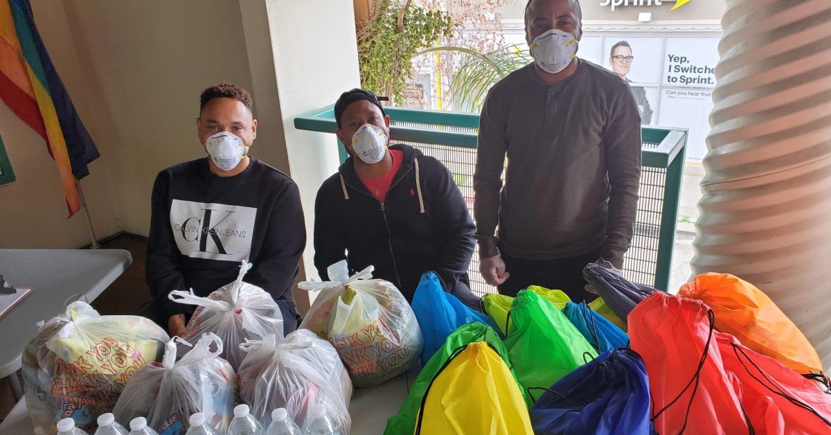 Three men at the Oakland LGBTQ Community Center, sitting behind a table with packaged food and other supplies.