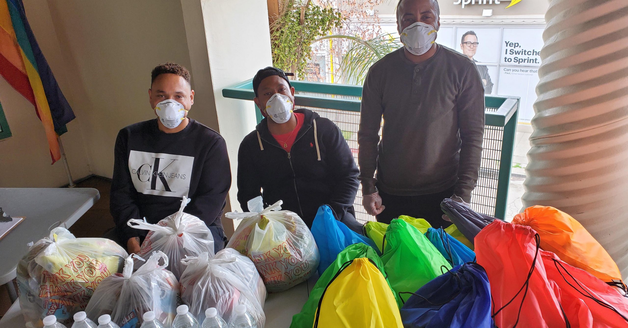 Three men at the Oakland LGBTQ Community Center, sitting behind a table with packaged food and other supplies.