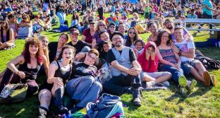 A color photograph of a group of about 20 individuals sitting together on some grass at Dolores Park in San Francisco during the Trans March of 2018. There are info booth tents and a crowd of people roaming in the background.