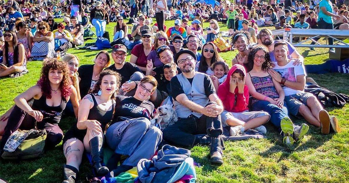 A color photograph of a group of about 20 individuals sitting together on some grass at Dolores Park in San Francisco during the Trans March of 2018. There are info booth tents and a crowd of people roaming in the background.