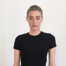 A caucasian woman wears a black shirt and has a shaved head.